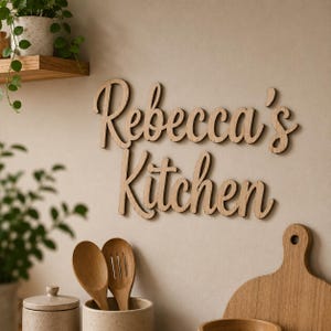 May include: Wooden sign reading "Rebecca's Kitchen" in a cursive script, mounted on a neutral wall. Kitchen utensils and decor are in the foreground. A wooden shelf with a potted plant is also visible in the image.