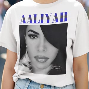 May include: White t-shirt featuring a black and white image of Aaliyah with the text "AALIYAH" in blue and black above the image. The text "LEGACY OF AALIYAH" is below the image. The model is wearing the shirt.