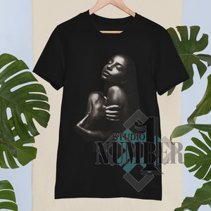 May include: Black t-shirt with a graphic of a woman with her eyes closed and arms wrapped around her body. The text "Studio Number 1" is printed in the bottom right corner of the shirt.