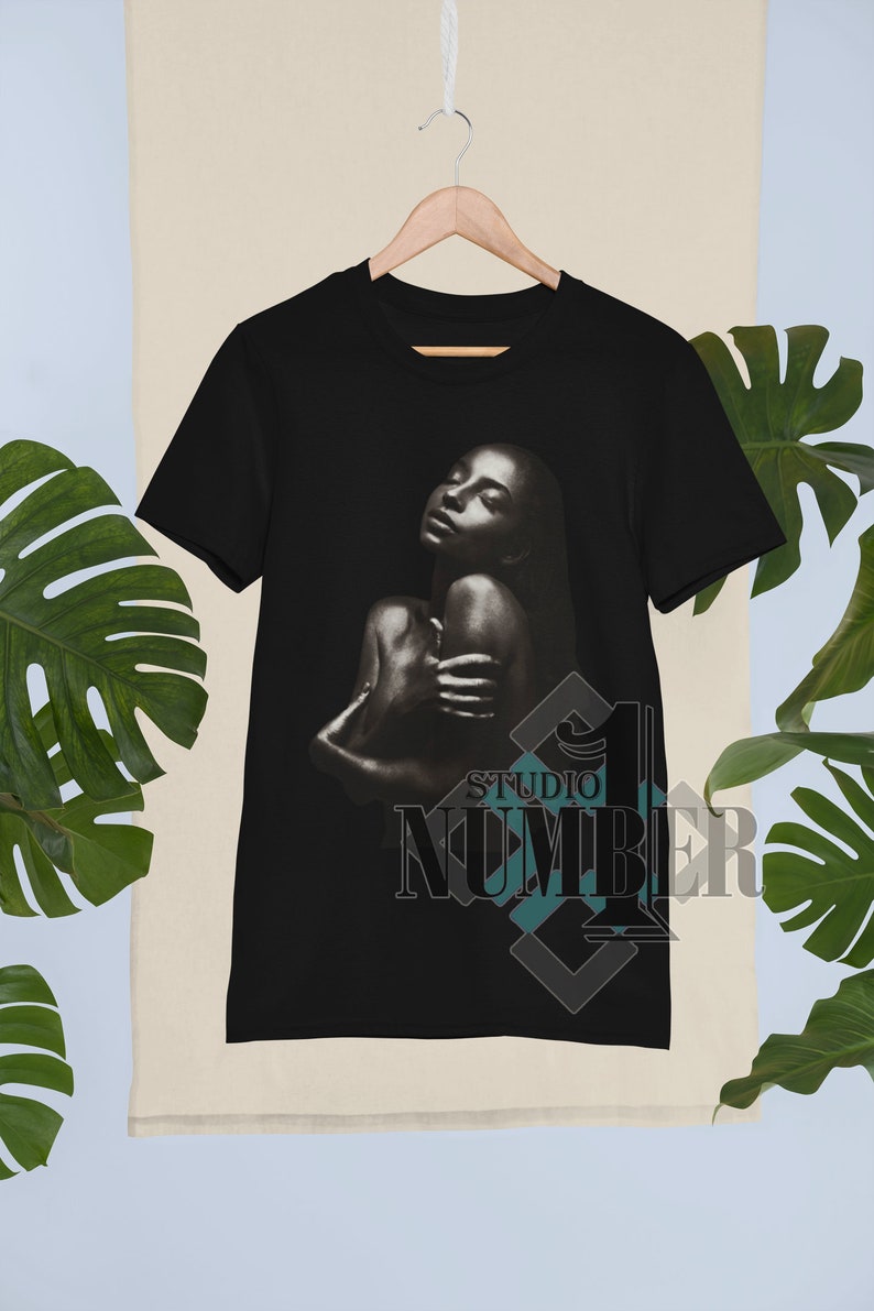 May include: Black t-shirt with a graphic of a woman with her eyes closed and arms wrapped around her body. The text "Studio Number 1" is printed in the bottom right corner of the shirt.