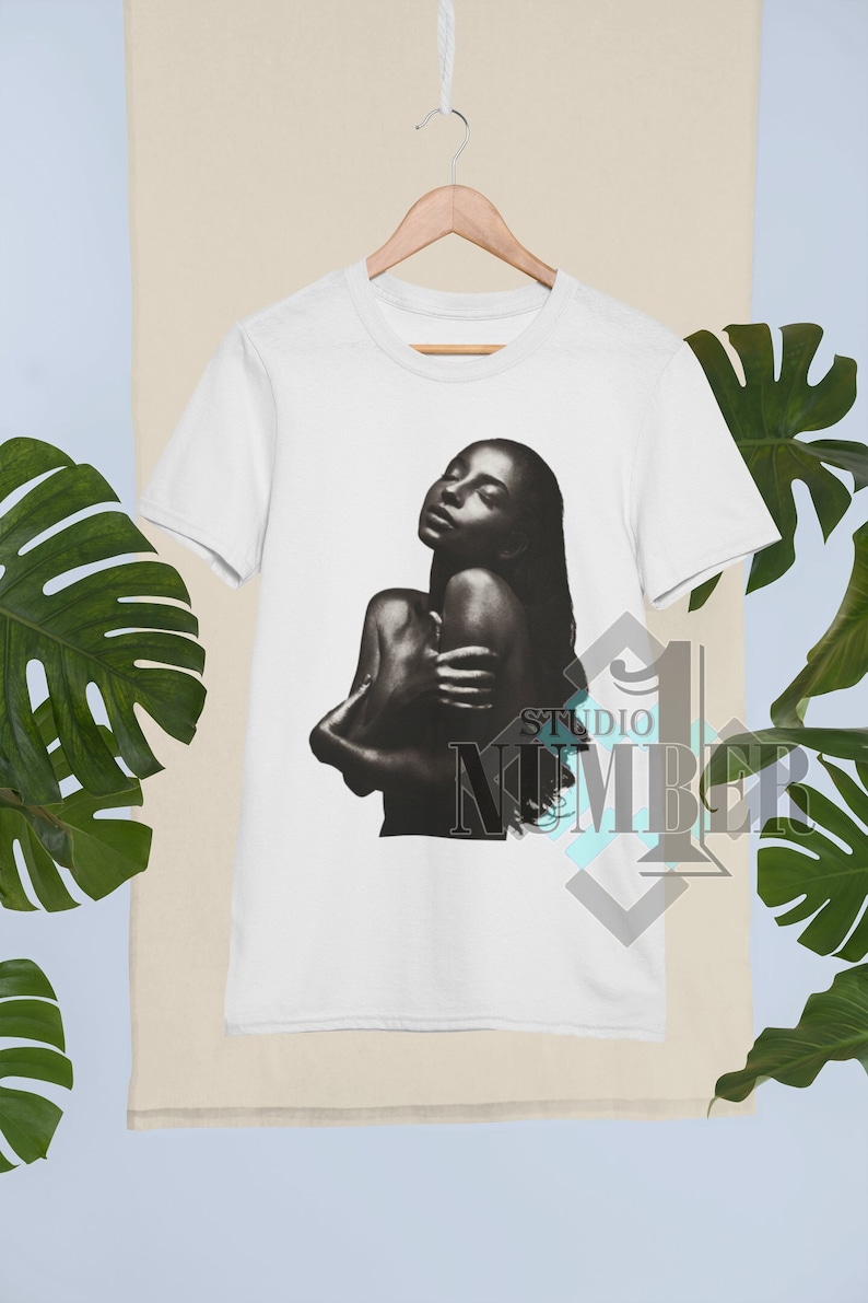 May include: White t-shirt with a black and white image of a woman with long hair, printed on the front. The image is of a woman with her arms wrapped around herself, looking up.