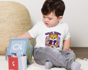 May include: A white toddler t-shirt with a purple and yellow cartoon tiger holding a football. The text "Geaux Tigers!" is printed on the shirt.