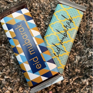 Op de afbeelding: Twee chocolade repen verpakt in folie met feestelijke ontwerpen. Eén reep heeft een blauw, goud en wit geometrisch patroon met de tekst "eid mubarak" in zwart. De andere reep heeft een turquoise en geel patroon met de tekst "eid mubarak" in zwart.