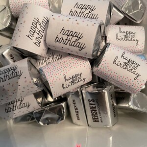 Könnte beinhalten: Eine weiße Schüssel gefüllt mit kleinen Schokoladenriegeln, die in Silberfolie verpackt sind. Jeder Riegel ist mit einer weißen Papierverpackung verziert, auf der der Text "happy birthday" in Schwarz gedruckt ist. Die Verpackungen haben ein Konfetti-Muster in Rosa, Blau und Grün.