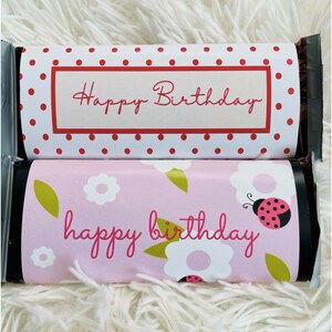 May include: Two chocolate bar wrappers with birthday greetings. The top wrapper is white with red polka dots and the text "Happy Birthday" in red. The bottom wrapper is pink with white flowers, green leaves, and a ladybug and the text "happy birthday" in pink.
