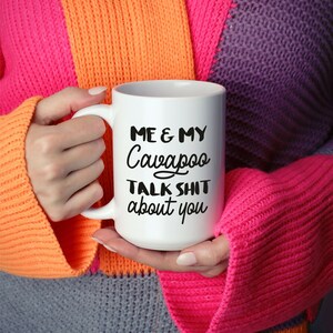 May include: White ceramic coffee mug with black text that reads "Me & My Cavapoo Talk Shit About You". The mug is being held by a person wearing a colourful sweater.