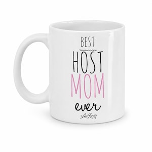 Pode incluir: Caneca de cerâmica branca com o texto "BEST HOST MOM ever" em letras pretas e rosa. A caneca tem uma alça curva e um pequeno ramo de folhas na parte inferior.