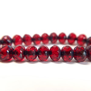 May include: A strand of faceted, deep red beads. The beads are evenly sized and have a glossy, reflective surface. The color is a rich, dark red, and the beads appear to be strung together. The background is plain white.