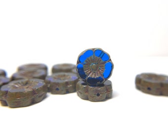 May include: Close-up of several flower-shaped beads. The beads are a dark, metallic color with a blue flower design in the center. The beads are arranged on a white surface.