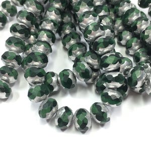 May include: Strands of faceted beads in silver and dark green. The beads have a metallic silver finish on the top and bottom, with a dark green, geometric pattern in the center. The beads are oval-shaped and appear to be strung together.