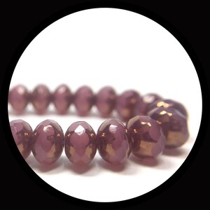 May include: A strand of round, faceted beads in a deep mauve color with a metallic, reflective finish. The beads are arranged in a horizontal line, showcasing their uniform shape and the light-catching properties of their polished, multi-faceted surfaces.