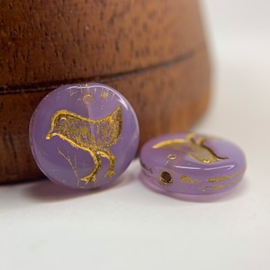 May include: Two purple glass beads with gold bird designs. The beads are round and have a hole in the center for stringing.