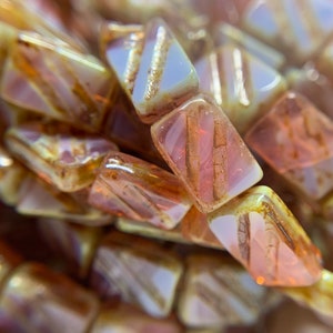 May include: A strand of glass beads with a mix of light brown, pink, and white colors. The beads have a textured, iridescent finish and are shaped like elongated rectangles.