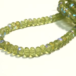 May include: A strand of green peridot beads with iridescent facets. The beads are faceted and have a slightly irregular shape.