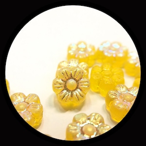 May include: A collection of small, translucent yellow flower-shaped beads with an iridescent finish. Each bead features a six-petaled design with a raised center, reflecting light with a subtle shimmer across the surface of the glass material.