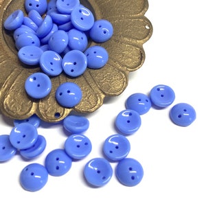 May include: A collection of light blue, round buttons with two holes each. The buttons are scattered on a white surface and in a decorative, antique gold-colored dish. The buttons have a glossy finish and are evenly spaced.