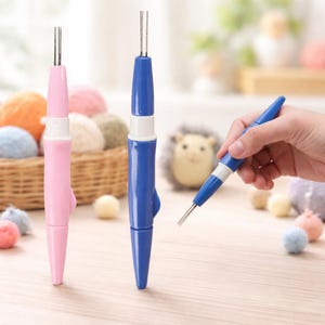 May include: Three punch needle tools in the image. Two are standing upright, one pink and one blue. The third is being held by a hand. The tools have a white section in the middle and a metal needle tip. The background includes yarn and a small hedgehog.