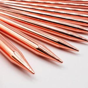 May include: A set of rose gold knitting needles with pointed tips. The needles are arranged in a fan shape, with the tips pointing towards the right side of the image.