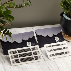 Op de afbeelding: Twee sets breinaalden en etuis. De etuis zijn zwart met een grijze geschulpte bovenkant en een schapenlogo. De naalden zijn zwart en zilver, vastgehouden met witte elastiekjes, en staan op een witte achtergrond.