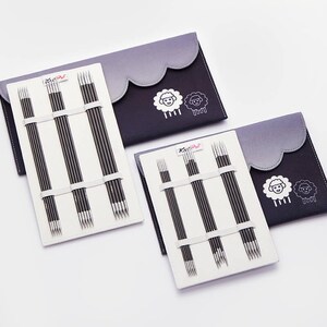 Op de afbeelding: Drie sets breinaalden in zwarte en witte etuis. Elke etui heeft een grijze geschulpte bovenkant en een schapenlogo. De naalden worden op hun plaats gehouden met witte elastische banden. De merknaam "KnitPro" is zichtbaar.