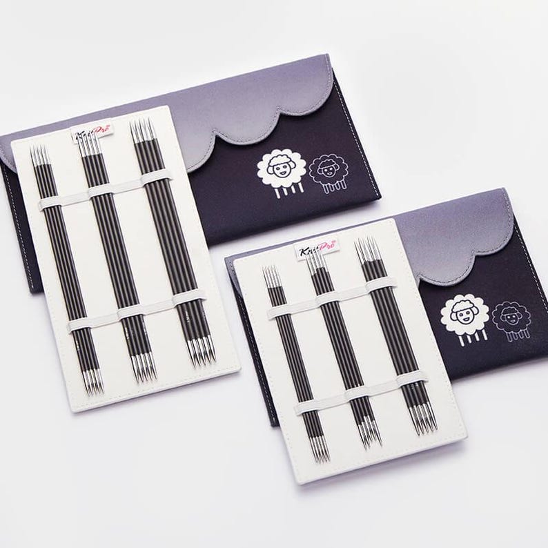 Op de afbeelding: Drie sets breinaalden in zwarte en witte etuis. Elke etui heeft een grijze geschulpte bovenkant en een schapenlogo. De naalden worden op hun plaats gehouden met witte elastische banden. De merknaam "KnitPro" is zichtbaar.