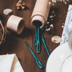 May include: A wooden box with a lid open, revealing several teal blue wooden sewing needles. The box is sitting on a wooden surface with a white fabric and a white ceramic saucer.
