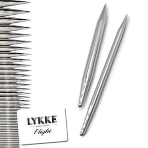 May include: Silver knitting needles with pointed ends, presented with a white card bearing the text "LYKKE - MAKE HAPPY - Flight". The image suggests a craft or hobby, with the needles set against a white backdrop.