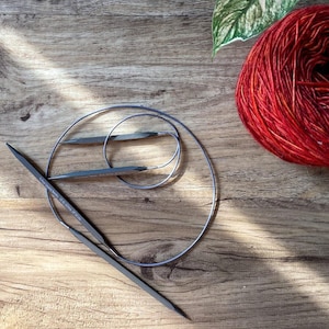 May include: A set of two circular knitting needles with a blue cord. The needles are made of metal and have a black finish. The needles are lying on a wooden surface.