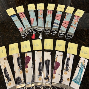 May include: A collection of sixteen fabric key fobs with various vintage dress designs. Each fob has a silver-colored metal ring and a coordinating tassel or charm. The key fobs are numbered from one to sixteen on yellow sticky notes.