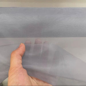以下が含まれることがあります： 滑らかな質感の薄いグレーのシアー生地。生地は手で持たれており、ドレープと透明度がわかります。