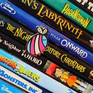 May include: A pink, white, and blue enamel pin of a bee with a black outline. The pin is sitting on a stack of DVD cases with movie titles visible, including "Paranorman", "Labyrinth", "Onward", "Before Christmas", "My Neighbor Totoro", "Monsters vs. Aliens", and "Monsters, Inc."