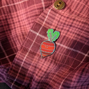 May include: A red and green enamel pin of a cartoon beet with a smiling face. The pin is attached to a red and white plaid shirt.