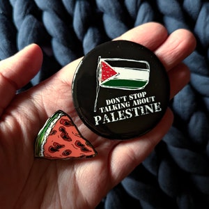 May include: Two pins: a black button with the Palestinian flag and the text "DON'T STOP TALKING ABOUT PALESTINE", and a watermelon slice pin with red, green, and black details. The pins are on a blue background.