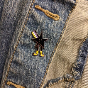 May include: A black star-shaped enamel pin with yellow and purple stripes, a smiling face, and yellow legs. The pin is attached to a denim jacket.