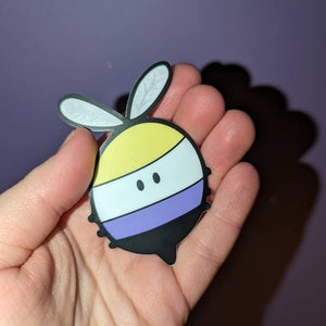 May include: A black and white bee sticker with yellow, white, and purple stripes. The bee has two small black eyes and a pair of white wings.