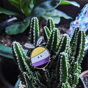 Puede incluir: Un pin de plata con forma de abeja de dibujos animados con rayas amarillas y negras y una raya morada y blanca en el medio. La abeja está sentada en un cactus verde.