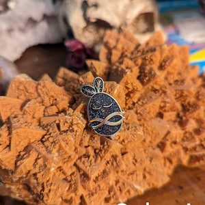 May include: A black enamel pin with a glittery finish, featuring a stylized sleeping creature with a white outline and rainbow accents. The pin is on a brown, textured surface. Color A.
