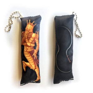 May include: Two black fabric keychains with different designs. One keychain features a gold-colored figure in armor, the other features a black whip.