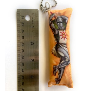 May include: A small, orange and yellow pillow with a printed image of a knight in armor. The knight is holding a sword and has a sun symbol on his chest. The pillow has a keychain attached to it.