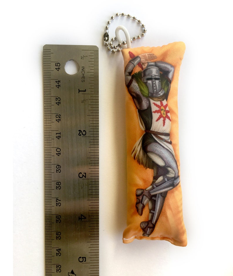 May include: A small, orange and yellow pillow with a printed image of a knight in armor. The knight is holding a sword and has a sun symbol on his chest. The pillow has a keychain attached to it.
