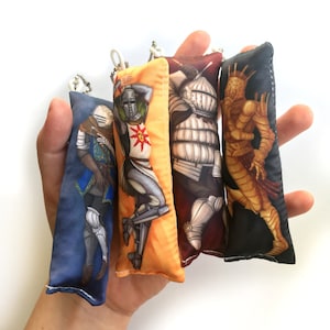May include: Four colorful fabric keychains featuring different characters from the video game Dark Souls. The keychains are made of soft fabric and have a plush texture. The characters include a knight in blue armor, a knight in gold armor, a knight in silver armor, and a creature with a crown.