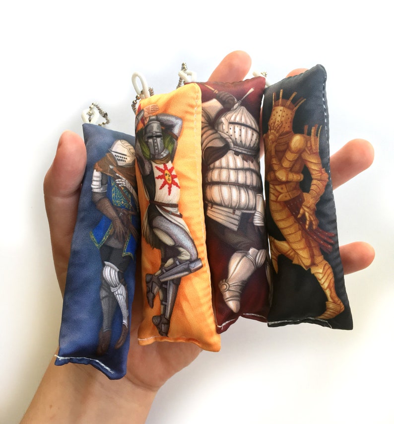 May include: Four colorful fabric keychains featuring different characters from the video game Dark Souls. The keychains are made of soft fabric and have a plush texture. The characters include a knight in blue armor, a knight in gold armor, a knight in silver armor, and a creature with a crown.