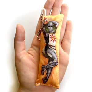 May include: A yellow and orange pillow keychain with a graphic of a knight in armor holding a sword and shield. The knight is wearing a helmet with a sun design on the front. The keychain has a white string loop and a silver chain.
