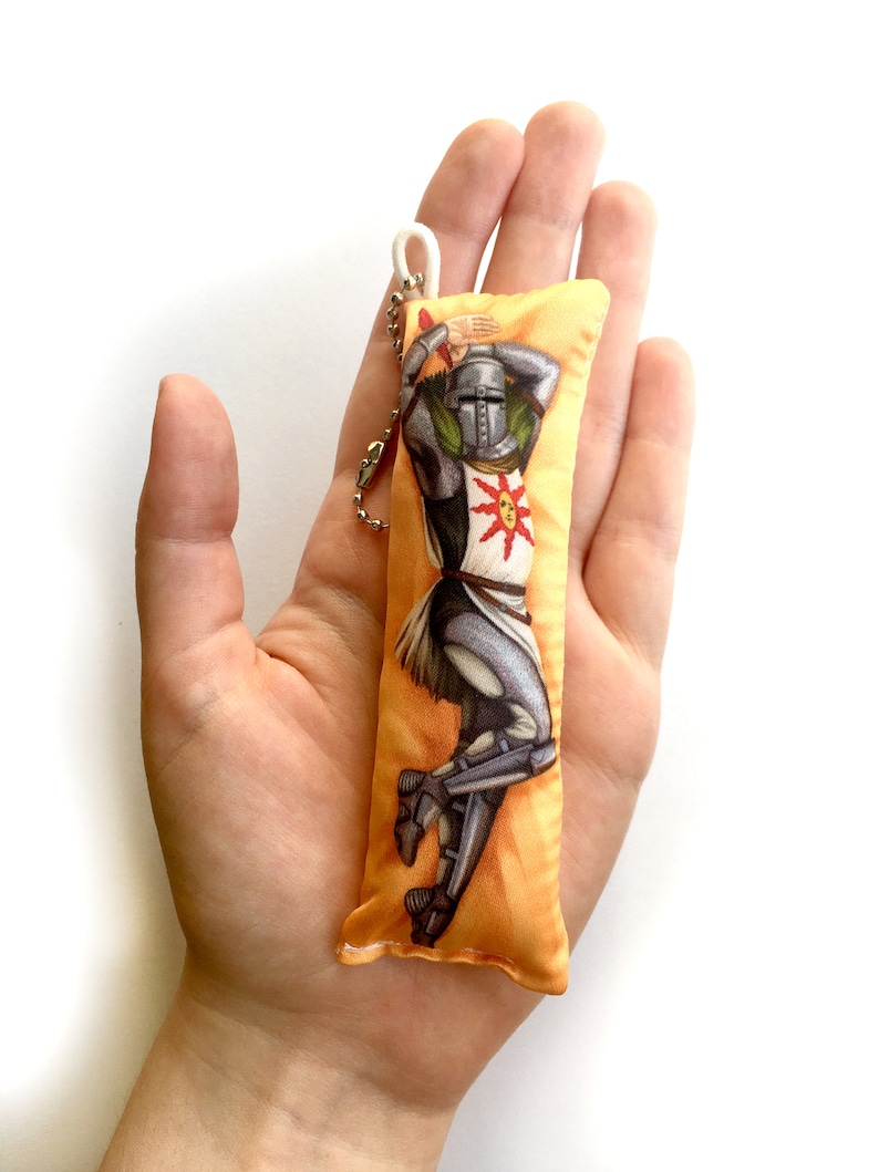 May include: A yellow and orange pillow keychain with a graphic of a knight in armor holding a sword and shield. The knight is wearing a helmet with a sun design on the front. The keychain has a white string loop and a silver chain.
