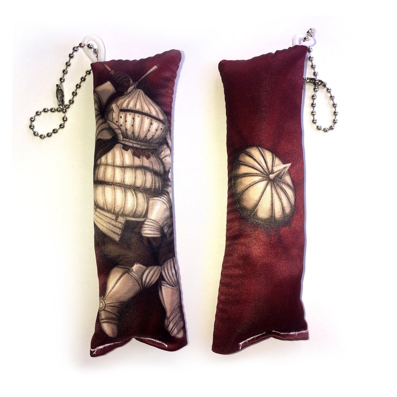 May include: Two red plush keychain pillows with a white chain. One pillow features a knight in armor, the other features a helmet.