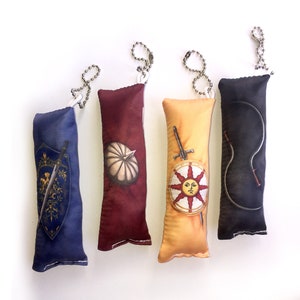 May include: Four plush keychain charms featuring different designs from the Dark Souls video game series. The charms are made of soft fabric and have a loop for attaching to a keychain. The designs include a shield with a sword, a sun with a face, a helmet, and a curved sword.