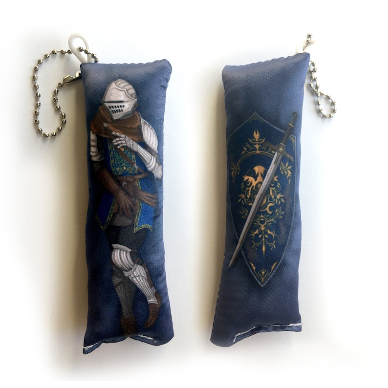May include: Two plush keychain pillows with a dark blue background. One pillow features a knight in blue and silver armor, while the other pillow features a shield with a sword and a golden eagle design.