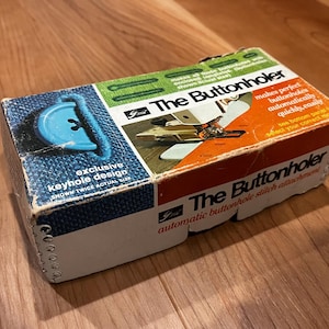 Könnte beinhalten: Ein rechteckiger Pappkarton für den The Buttonholer Knopfloch-Nähaufsatz. Die Verpackung hat blaue, grüne und orangefarbene Felder mit Text, darunter "exclusive keyhole design" und "makes perfect buttonholes automatically".