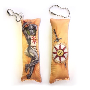 May include: Two plush keychain pillows featuring a knight in armor and a shield with a sun design. The knight is wearing a helmet and holding a sword. The shield has a sun with a face on it. Both pillows are beige with a gold trim.