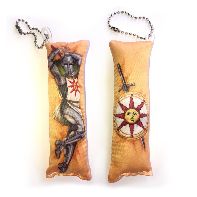 May include: Two plush keychain pillows featuring a knight in armor and a shield with a sun design. The knight is wearing a helmet and holding a sword. The shield has a sun with a face on it. Both pillows are beige with a gold trim.